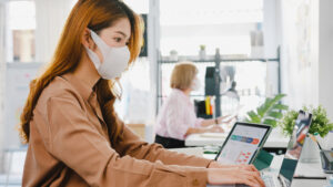 asia businesswoman entrepreneur wearing medical face mask social distancing new normal situation virus prevention while using laptop back work office